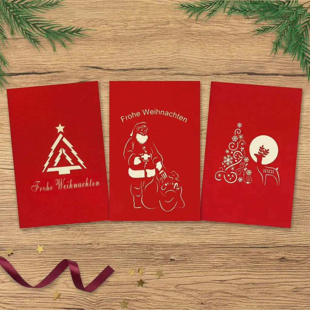 Drei rote Weihnachtskarten mit modernem Tannenbaum, Weihnachtsmann mit Gabensack und Rentier neben einem verzierten Baum auf einem hellen Holztisch