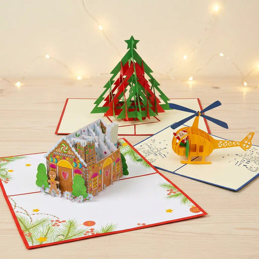 Drei geöffnete LINPopUp Pop-Up-Weihnachtskarten mit Lebkuchenhaus auf weihnachtlich bedruckter Kartenfläche, rot-grünem Tannenbaum und gelbem Weihnachtsmann-Helikopter