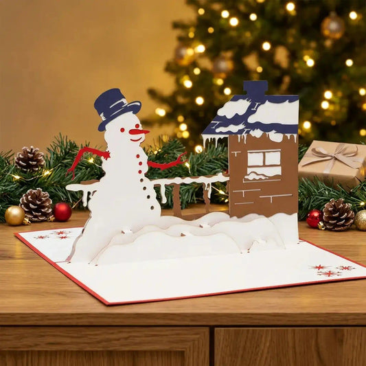 Offene Schneemann-am-Haus-Pop-Up-Karte auf einem Sideboard mit Tannengrün, Weihnachtskugeln und warmen Lichtern