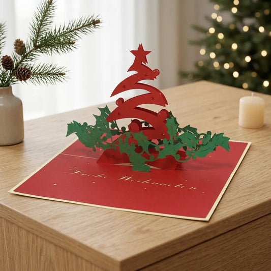 Geöffnete rote Pop-Up-Weihnachtskarte mit geschwungenem Tannenbaum und grünen Blattzweigen auf einem Eichensideboard, mit Tannenzweigen, Kerze und Weihnachtslichtern im Hintergrund.