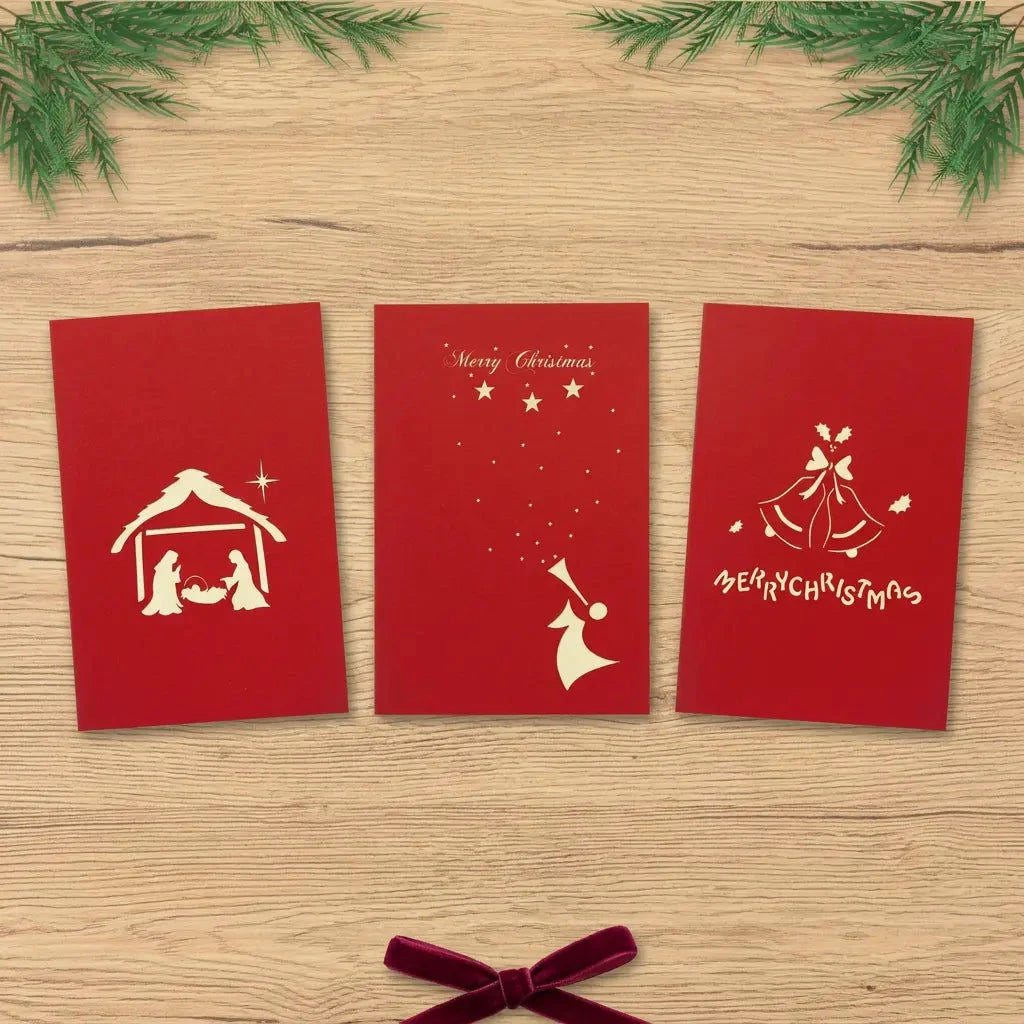 Drei rote Weihnachtskarten mit Krippe, Engel und Glocken nebeneinander auf einem hellen Holztisch mit Tannenzweigen