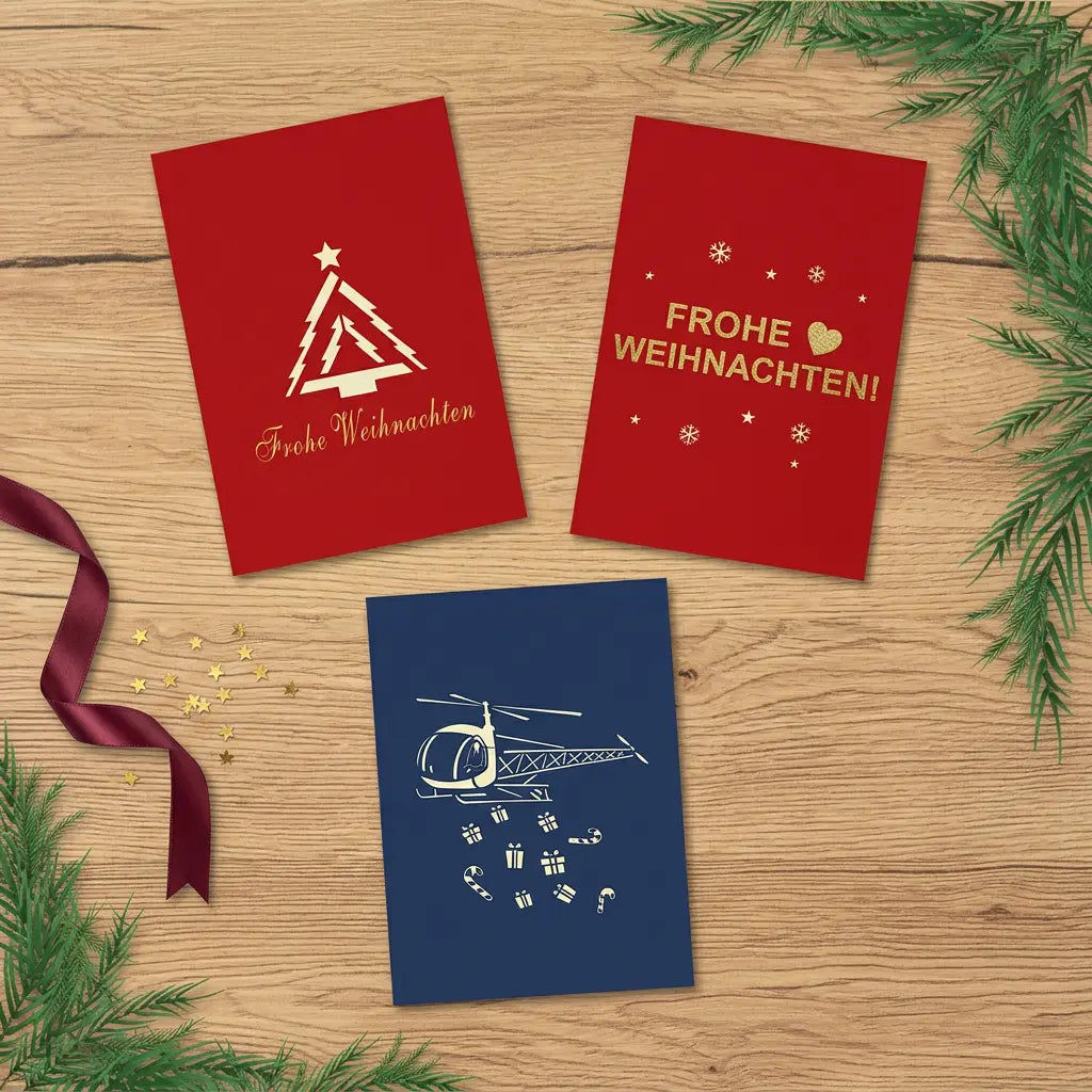 Drei geschlossene Weihnachtskarten mit modernem Tannenbaum, goldenem Weihnachtsgruß und Helikoptermotiv auf einem festlich dekorierten Holztisch