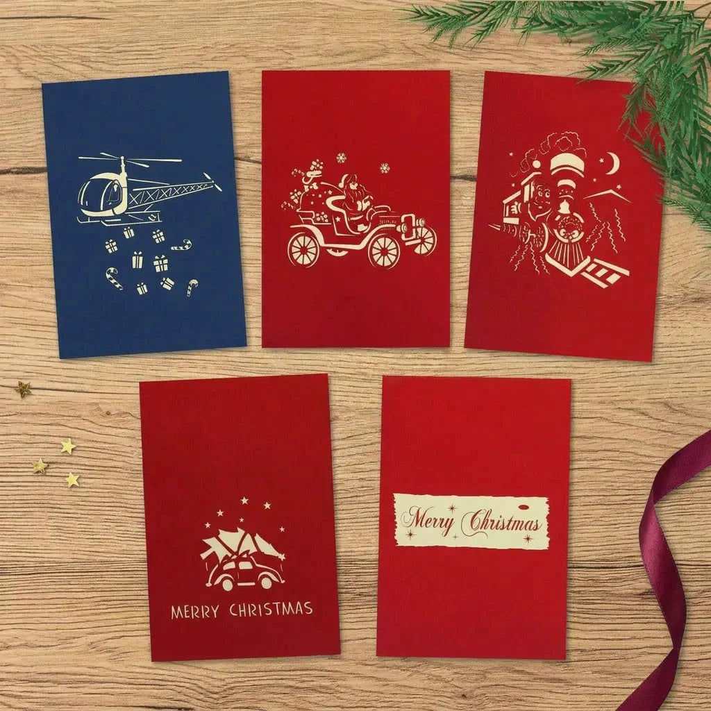 Fünf geschlossene Weihnachtskarten mit Helikopter, Oldtimer, Weihnachtszug, Auto mit Tannenbaum und Weihnachtsgruß auf einem hellen Holztisch.