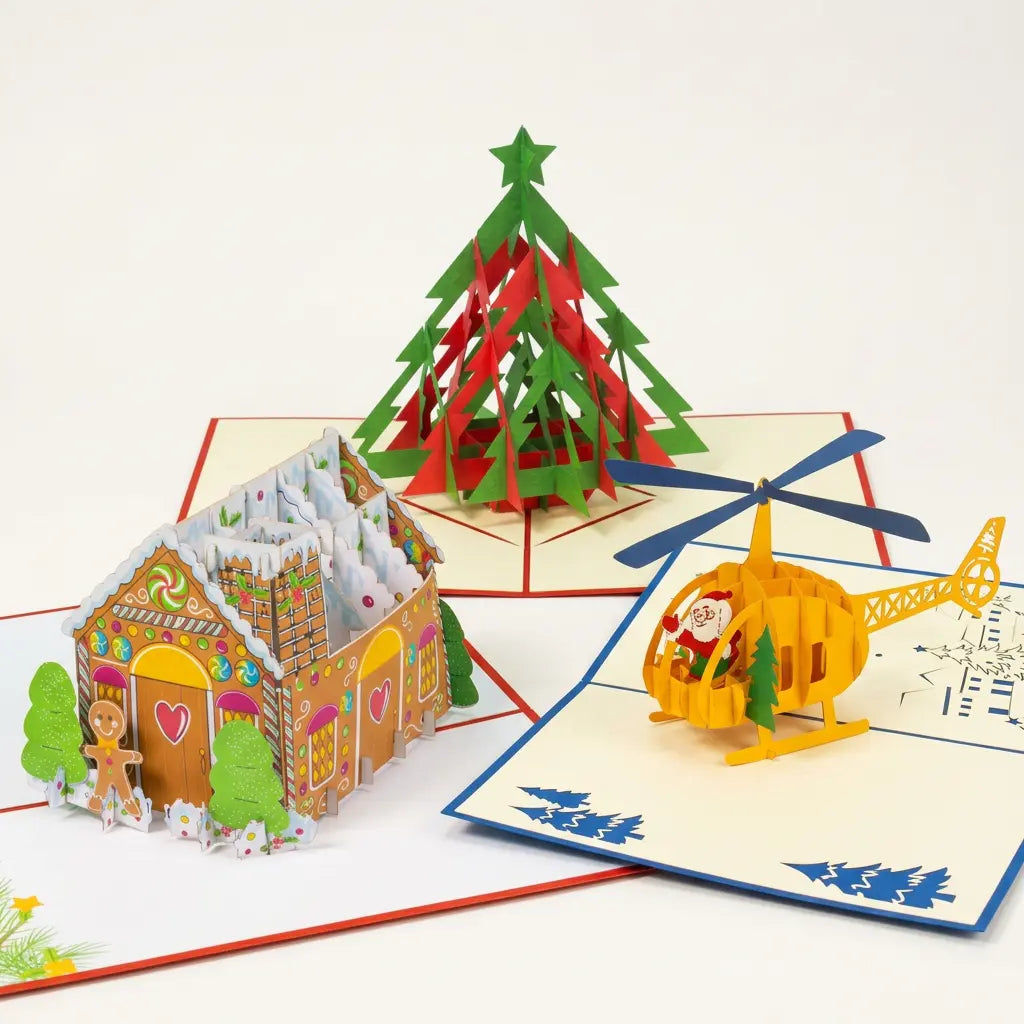 Drei geöffnete LINPopUp Pop-Up-Weihnachtskarten mit Lebkuchenhaus, rot-grünem Tannenbaum und gelbem Weihnachtsmann-Helikopter auf hellem Hintergrund