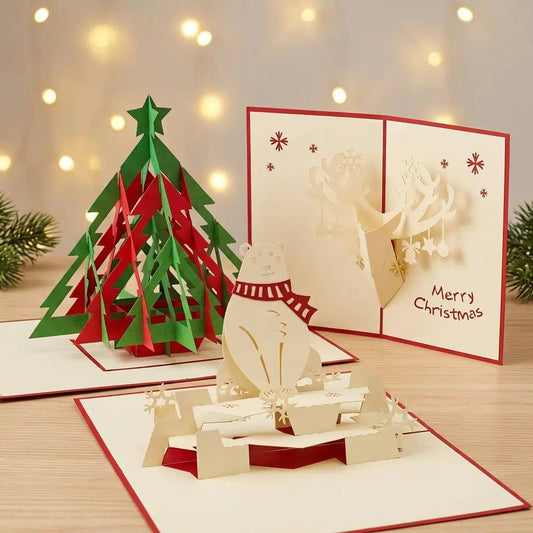 Drei geöffnete LINPopUp Pop-Up-Weihnachtskarten mit Tannenbaum, Eisbär mit rotem Schal und geschmücktem Rentierkopf auf hellem Holz vor warmen Weihnachtslichtern