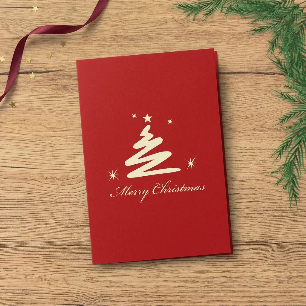 Geschlossene rote Weihnachtskarte mit cremefarbenem, geschwungenem Tannenbaum, Sternen und „Merry Christmas“ auf einem festlich dekorierten Eichentisch.